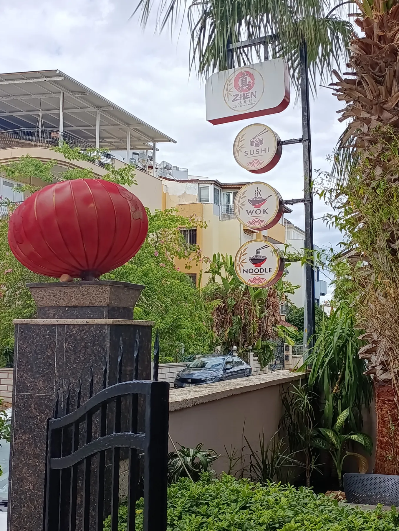 Kapı direğindeki kırmızı fener ve arkada Zhen, Sushi, Wok, Noodle tabelası